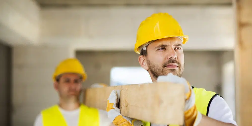 Zwei Männer tragen einen Holzbalken, sie wollen ihr Haus selber bauen